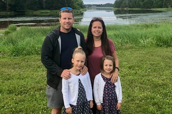 Carrie Nelson, a middle-aged woman, smiling with her husband and two young daughters in a field.